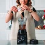 A woman pours a sachet into a blender in a festive kitchen setting.