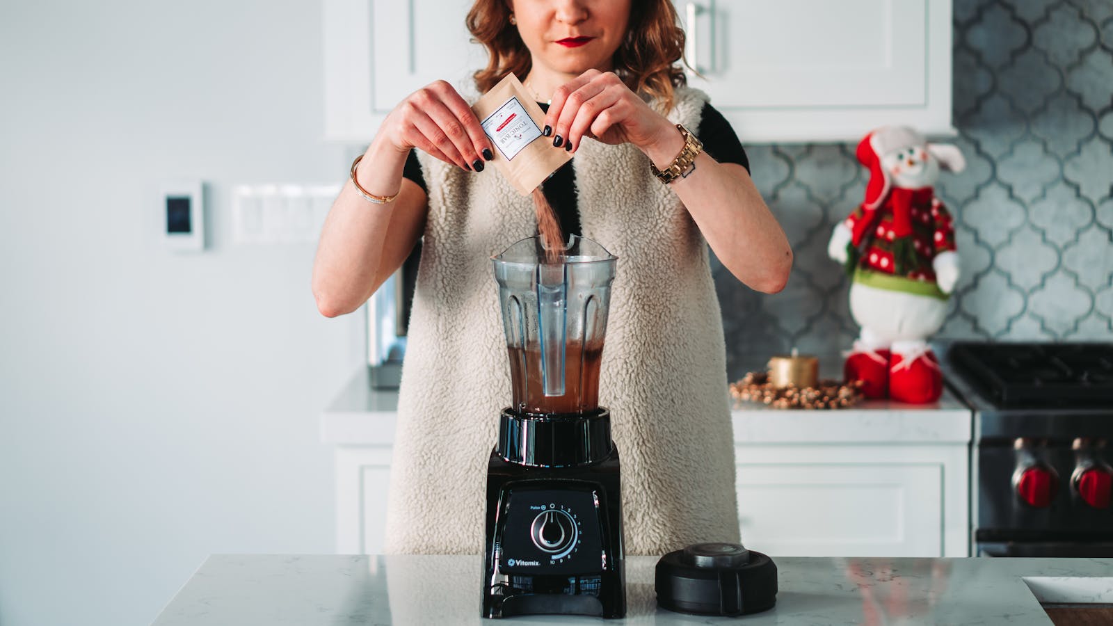 A woman pours a sachet into a blender in a festive kitchen setting.