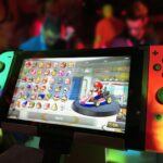 Close-up of a Nintendo Switch showing Mario Kart selection screen, gaming atmosphere in the background.