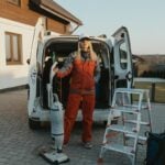 A cleaner in uniform stands beside a van with cleaning tools outside a modern house at sunset.
