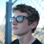 a young man wearing sunglasses standing next to a fence